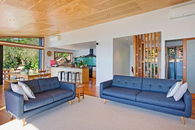 Bright open-plan living area with two blue sofas, a light neutral rug, and timber flooring. The room features a warm timber-lined ceiling with integrated ventilation grilles and recessed lights, white walls, large windows, and an open kitchen with a timber island and dark rangehood. A vertical timber screen partially separates the living room from the entry or adjacent area. The overall design is clean, contemporary, and connected to greenery outside.