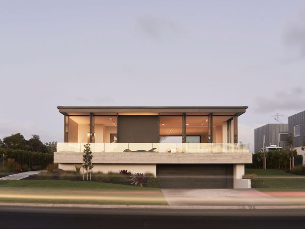 Street-facing exterior view of a contemporary multi-level residence with a broad flat roof, extensive upper-level glazing, a deep balcony terrace with a translucent balustrade, and a recessed double garage beneath. The facade combines warm neutral rendered surfaces with dark slender framing and a dark garage door, set within a landscaped suburban frontage.