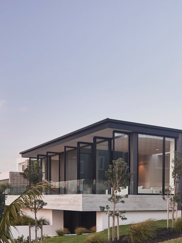 Exterior view of a contemporary two-storey residence with a low-pitched roof, extensive floor-to-ceiling glazing, dark metal window and door frames, and a cantilevered upper level over a recessed lower area. The home includes a glass balustrade, pale masonry or stone-clad walls, landscaped planting, and a clean indoor-outdoor connection.
