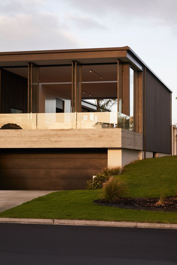 Exterior view of a contemporary single-storey home on a gently sloped landscaped site. The building features a low-pitched roof, dark vertical cladding, extensive glazed openings, warm timber framing, a translucent balcony balustrade, and a large dark timber garage door. The home is photographed from the street at dusk or in soft evening light.