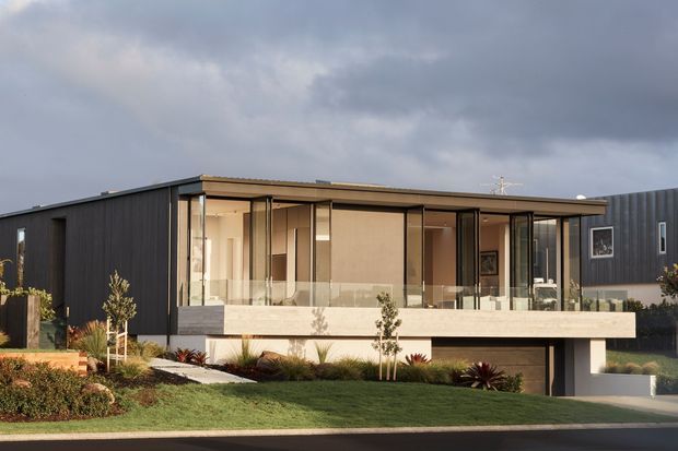 Exterior photograph of a contemporary flat-roofed, multi-level residence. The home features dark vertical cladding, extensive floor-to-ceiling glazing, a projecting concrete balcony or terrace with glass balustrades, and an integrated lower-level garage. Minimal landscaped planting and a sloping front lawn surround the building.