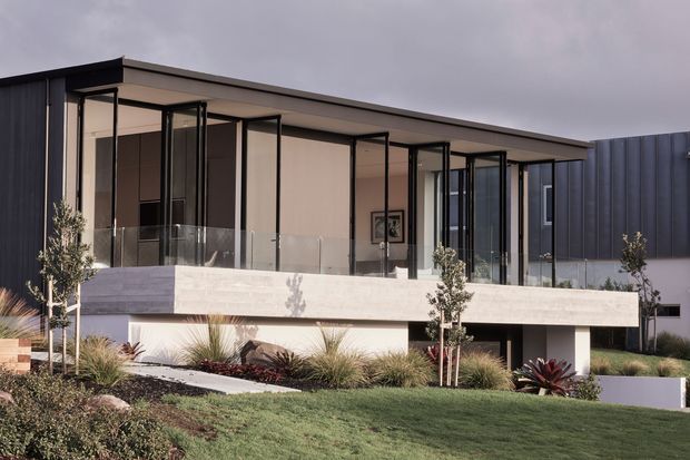 Exterior view of a contemporary single-storey residence with a broad flat roof, expansive floor-to-ceiling glazing, slender dark window frames, a projecting concrete balcony or terrace slab, and dark vertical cladding. The home is set above a landscaped sloping lawn with ornamental grasses and young trees under an overcast sky.
