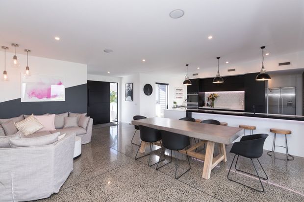 Open-plan contemporary kitchen, dining and living area with polished terrazzo-style flooring, a long timber dining table, upholstered seating, dark kitchen cabinetry, a large island bench, pendant lighting and integrated stainless-steel appliances. The palette is predominantly white, grey and black with warm timber accents.
