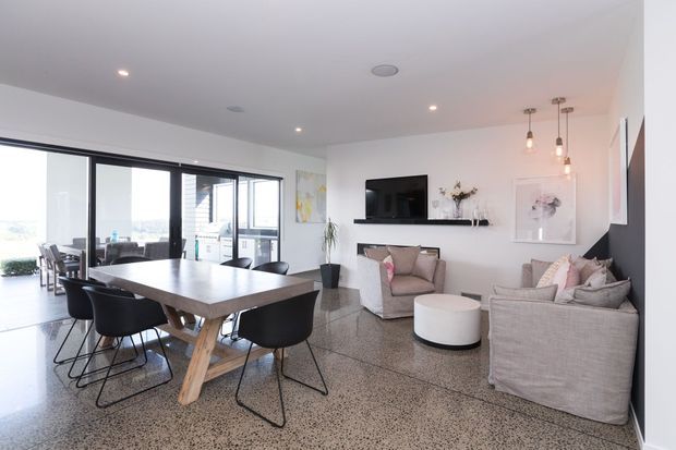 Bright open-plan living and dining area with polished terrazzo flooring, a large timber dining table with black chairs, a neutral fabric sofa and armchairs, wall-mounted television, pendant lights, and extensive black-framed glazing opening toward an outdoor balcony or terrace.