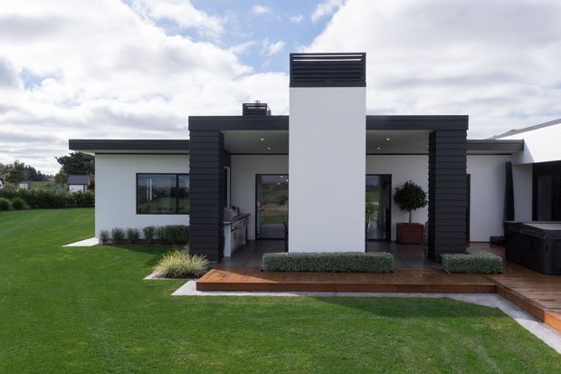 Single-storey modern house exterior with a flat roof, white rendered walls, dark horizontal cladding framing the central entry tower, and a timber deck running along the facade. The landscaped front lawn and simple planted beds complement the crisp, geometric facade.