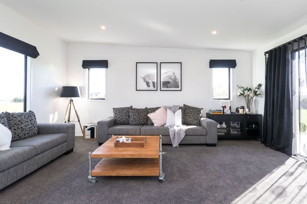 Bright contemporary living room with grey sofas, patterned cushions, a timber coffee table on casters, dark media console, framed artwork, floor lamp, and full-height dark curtains. White walls and ceiling, grey carpet, and abundant natural light create a comfortable, refined setting.
