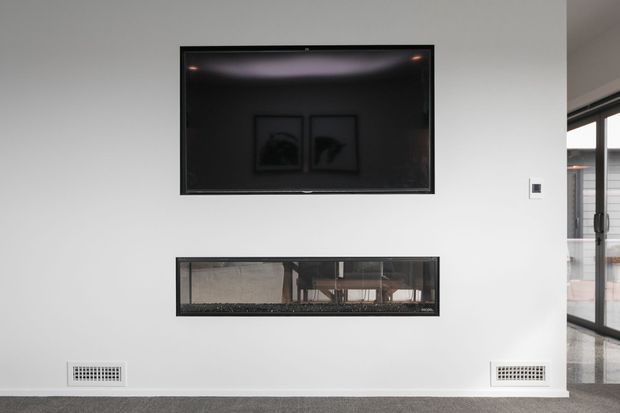 A minimalist white interior wall featuring a wall-mounted flat-screen television above a long, recessed glass-fronted fireplace or feature opening. Two low ventilation grilles sit near the floor, with dark carpet underfoot and floor-to-ceiling glazing visible at the right edge.