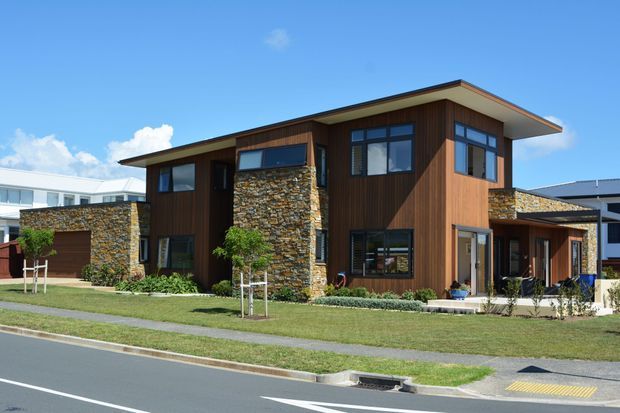 Exterior view of a contemporary multi-level residential building with warm vertical timber cladding, prominent rough stacked-stone feature walls, broad roof overhangs, and extensive dark-framed glazing. The landscaped front setback includes lawn, young trees, shrubs, a footpath and street frontage.