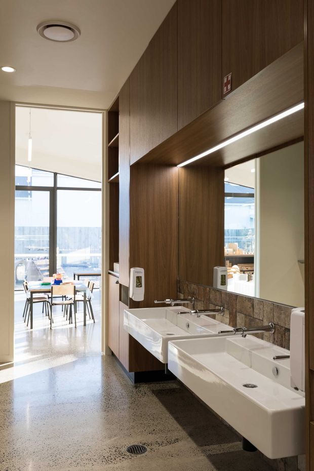 Contemporary commercial washroom with a row of wall-mounted white basins, chrome taps, a large mirror, and dark timber vanity cabinetry. A mottled stone or terrazzo-like floor, patterned stone splashback, integrated under-cabinet lighting, and full-height glazing connect the washroom to a bright communal dining or work area beyond.