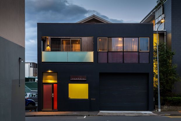 Street-facing view of a compact contemporary multi-storey residence at dusk, with a dark charcoal facade, projecting upper-level balcony with frosted glass balustrade, large glazed windows, a vivid yellow entry panel, red front door, and a wide black roller garage door. Neighbouring urban buildings and a narrow street frame the property.