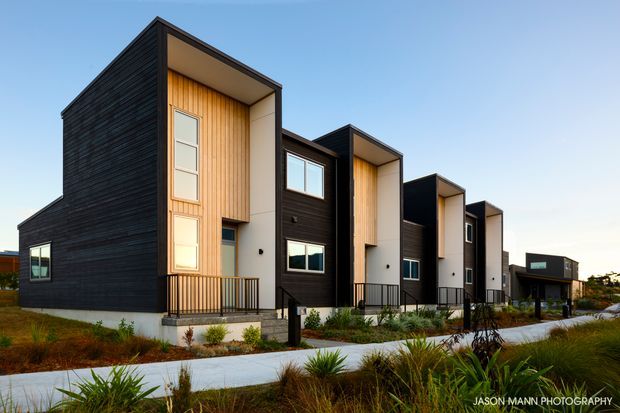Exterior architectural photograph of a row of contemporary multi-storey townhouses. The buildings feature dark horizontal cladding, warm vertical timber-panelled recessed entries, projecting box-like upper forms, flat rooflines, large rectangular windows, and landscaped native-style planting along a pedestrian path.
