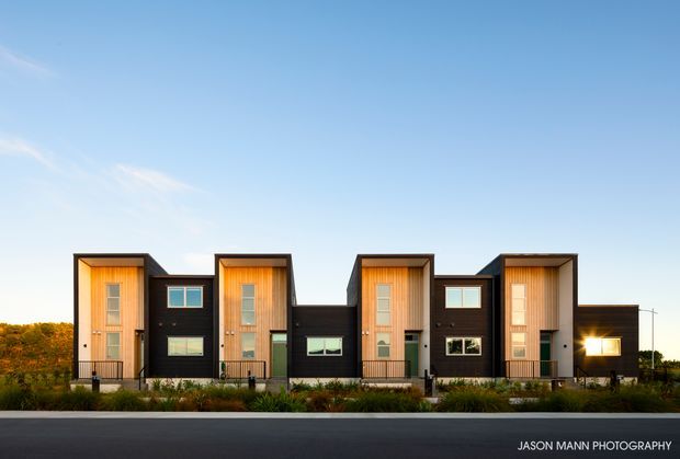 Front exterior view of a contemporary multi-unit residential building with a rhythmic row of tall projecting bays, dark cladding, warm vertical timber-lined entry recesses, large windows, and a flat roofline. The building sits behind landscaped planting and a paved street under a clear sky.