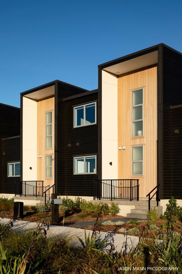 Exterior architectural photograph of a contemporary multi-unit residential building with repeated projecting entry bays, dark horizontal cladding, pale vertical timber-look feature panels, large windows, recessed soffits, and black metal railings. Low-maintenance landscaped planting and concrete paths occupy the foreground under clear blue sky.