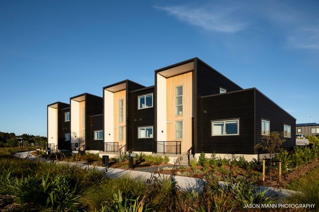 Exterior photograph of a contemporary multi-unit residential development with repeated tall, box-like entry volumes, dark horizontal cladding, warm timber-toned recessed facades, and large vertical windows. The buildings sit along landscaped pedestrian paths with native-style planting under a clear blue sky.