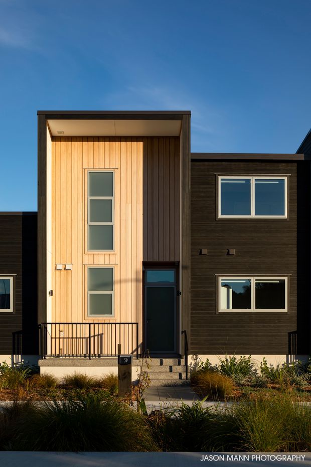 Front exterior view of a contemporary multi-storey townhouse or residential building. The facade combines dark horizontal cladding with a projecting entry volume clad in warm vertical timber boards, large rectangular windows, a recessed front door, and a simple black metal entry railing. Drought-tolerant ornamental grasses and low planting line the front path.