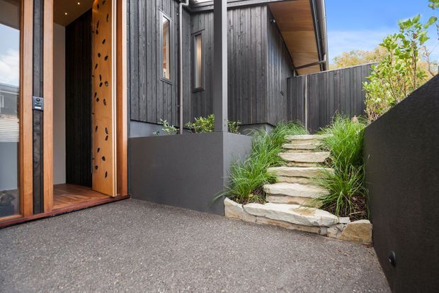 Exterior view of a contemporary dark-clad residential entry. A timber-framed glazed front door with decorative perforated timber panel opens onto a textured paved forecourt, while rough natural stone steps edged with ornamental grasses lead upward beside dark vertical cladding and garden walls.