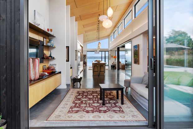 Bright open-plan contemporary home interior viewed through a large glazed opening, with a double-height living and dining area, exposed warm timber ceiling, polished concrete flooring, built-in timber shelving and cabinetry, pendant lights, a patterned rug, and full-height glazing overlooking a pool and outdoor setting.