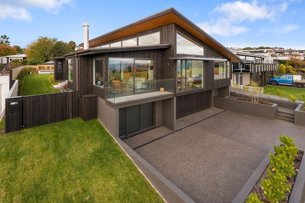 Exterior view of a contemporary two-storey home on a suburban site, featuring dark vertical timber cladding, a prominent pitched gable roof with warm timber soffits, extensive glazing, a projecting upper-level balcony, and a broad paved driveway with landscaped lawn.