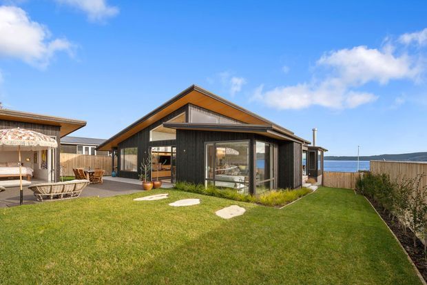 Exterior view of a modern single-storey home with dark vertical cladding, warm timber soffits, large glazed openings and a pitched gable roof. The building sits beside a neatly landscaped lawn and deck area, with a visible body of water beyond the fence. Outdoor seating and a large parasol occupy the left side of the yard.