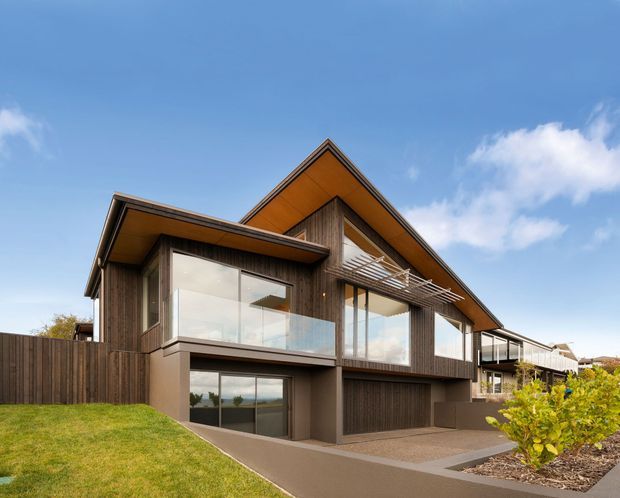 Exterior view of a contemporary multi-level residence with a steep asymmetrical gabled roof, dark vertical timber cladding, extensive glazing, and warm timber-lined soffits. The house includes projecting upper-level balconies with glass balustrades, an integrated lower-level garage, paved access, and landscaped lawn and planting.
