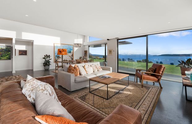Bright open-plan living room with floor-to-ceiling glazing overlooking a waterfront landscape. The space features white walls, polished concrete flooring, a large patterned rug, a brown leather sectional sofa, a light upholstered sofa, timber coffee tables, armchairs, indoor plants, artwork, and a dining area beyond.