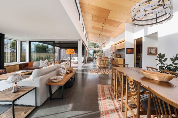 Bright open-plan residential interior combining a living room, dining area, and kitchen. The space features a long timber-clad ceiling, extensive black-framed glazing, polished dark flooring, white walls, warm timber joinery, a large timber dining table, and a sculptural chandelier. The living area includes a light sectional sofa and built-in window seating, while the kitchen and island are visible toward the rear.