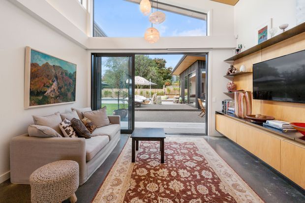 Bright contemporary living room with a double-height ceiling and clerestory glazing, opening through large sliding glass doors to a landscaped outdoor entertaining area. The room features a neutral upholstered sofa, patterned rug, low coffee table, integrated timber entertainment cabinetry with open shelving, and a wall-mounted television. Pendant lights hang in the tall void, while a large artwork adds colour.