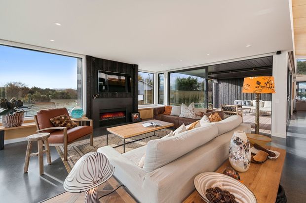 Bright contemporary living room with expansive floor-to-ceiling glazing overlooking a landscaped outdoor setting. The room features a large cream sectional sofa, leather lounge chairs, a timber coffee table, a dark stone or tile fireplace wall with a built-in fire, and warm timber flooring. An adjoining covered outdoor area is visible through the glazing.