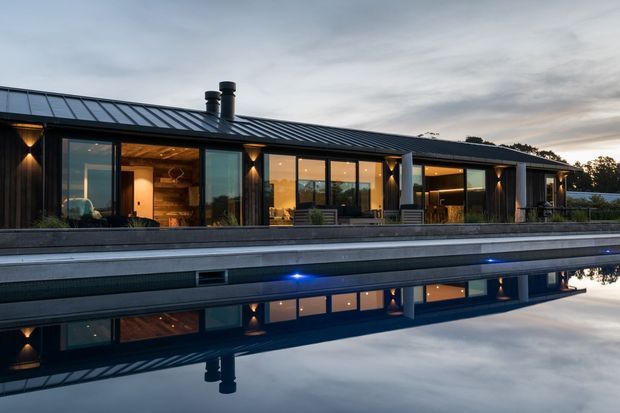 A long, low-slung contemporary residence viewed from across a reflecting swimming pool at dusk. The home features dark vertical cladding, extensive floor-to-ceiling glazing, a standing-seam metal pitched roof, warm interior lighting, and a broad pool terrace. The pool creates a strong mirror reflection of the facade and roofline.