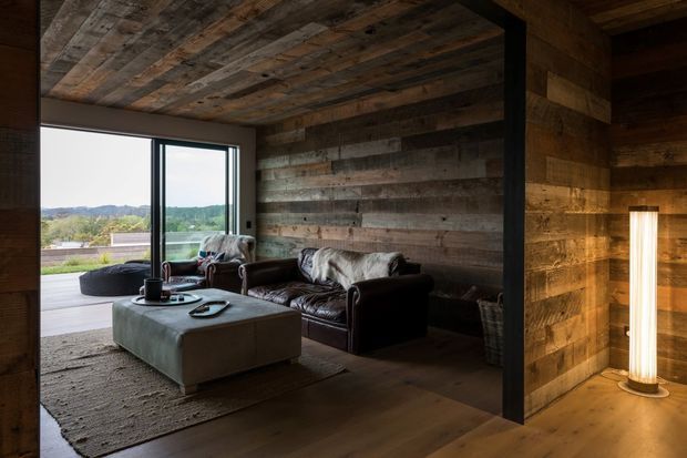 Rustic living room with dark leather sofas, a large upholstered ottoman, and a textured rug, enclosed by reclaimed timber wall and ceiling cladding. Full-height glazing opens toward a landscape view, while a warm vertical feature lamp adds ambient light beside a dark structural post.
