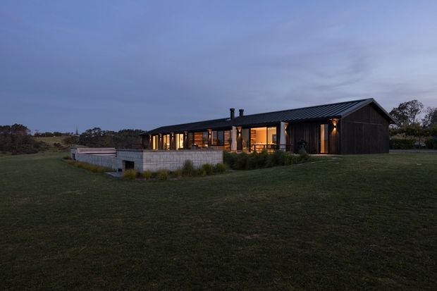 A single-storey contemporary rural house photographed at dusk, with a long low profile, dark vertical cladding, a dark pitched metal roof, extensive warm-lit glazing, and a small concrete terrace or retaining structure set within open grassed grounds.