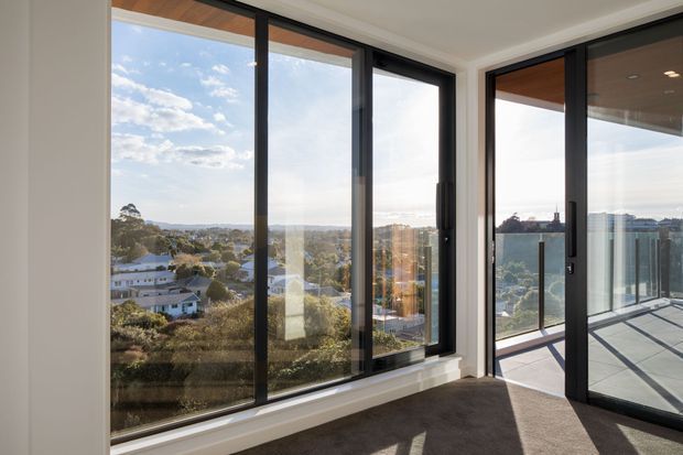 Bright contemporary interior corner with expansive floor-to-ceiling black-framed glazing and a glass door opening onto a balcony or terrace. The room has neutral white walls, a dark carpeted floor, and a timber-lined exterior soffit visible beyond the glazing. A suburban landscape and distant water or horizon are visible outside.