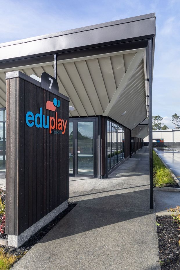 Exterior architectural photograph of a contemporary childcare centre entrance. The building features dark vertical timber-look cladding, extensive floor-to-ceiling glazing, a broad projecting roof canopy with a white exposed-panel soffit, black structural posts and framed doors, and landscaped planting along a paved walkway. The visible “eduplay childcare” signage identifies the commercial childcare facility.