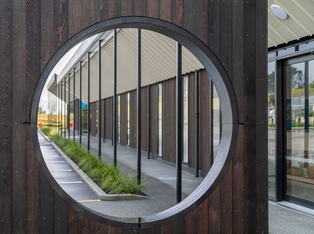 Close-up architectural exterior detail featuring a large circular opening or window set into dark vertical timber cladding. The opening frames a reflective view of a long glazed building frontage with slender dark frames, a covered walkway, concrete paving, and linear planting. Part of a pale soffit and adjacent glazed entry are visible at the right.