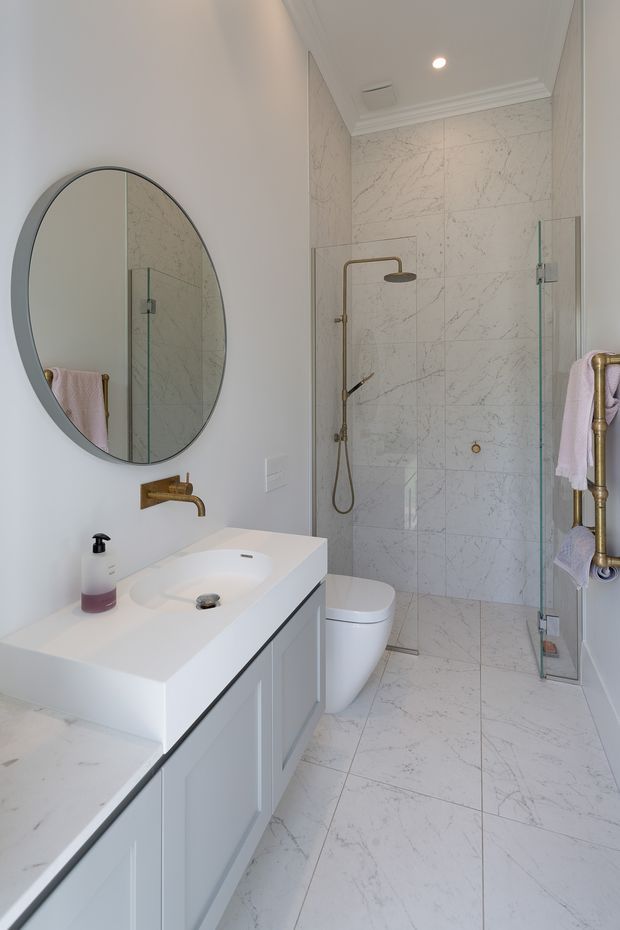 Bright, narrow contemporary bathroom with a white vanity and integrated basin, large round mirror, toilet, and walk-in glass shower enclosure. The walls and floor are clad in white marble-look veined tiles, with brushed brass shower fittings and heated towel rail accents. Recessed lighting and white decorative ceiling moulding create a clean, refined finish.