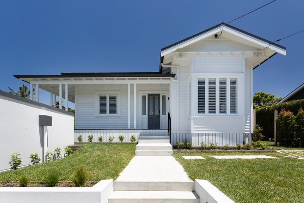 Front exterior view of a renovated or newly built white weatherboard house with a prominent gabled front wing, decorative eaves, white plantation shutters, covered entry porch, and a low white boundary wall. A concrete path and steps lead through a landscaped front lawn to the dark entry door. Clear blue sky and neighbouring residential context are visible.