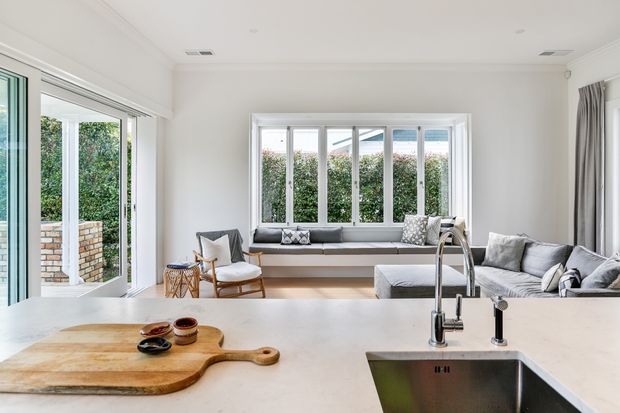 Bright open-plan kitchen and living area with a pale stone island benchtop, integrated stainless-steel sink and brushed metal tapware in the foreground. Beyond the island is a light, relaxed living space with a grey sectional sofa, window seat, and large white-framed windows opening to a leafy outdoor area and brick privacy wall. The room has white walls, white trim, light timber accents, and abundant natural light.
