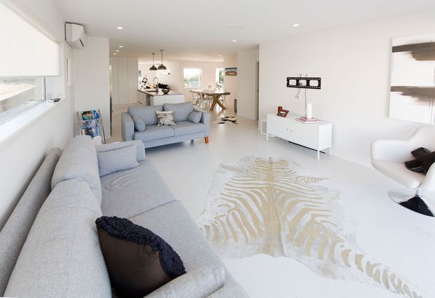 Bright, open-plan contemporary living area with pale grey sofas, a white media console, sculptural patterned rug, and minimal white furnishings. The living room opens onto a dining area and kitchen in the background, with abundant natural light and a clean, airy palette.