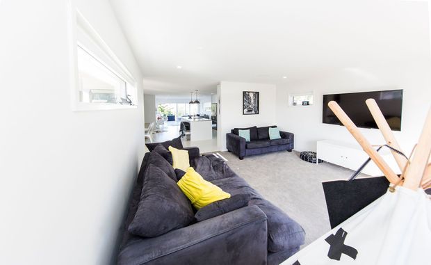 Bright open-plan living room with large dark sectional sofas, yellow accent cushions, a grey carpet, wall-mounted television, and white low-profile media cabinetry. The kitchen and dining area are visible beyond the living space, while a children's teepee occupies the foreground.