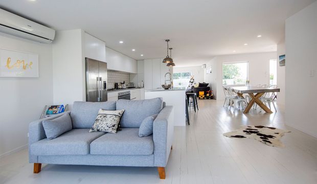 Bright open-plan residential interior with a light blue sofa in the foreground, a modern white kitchen with stainless-steel appliances, a dining area, and a small living or breakfast nook. The space has white walls and cabinetry, pale timber flooring, recessed lighting, and pendant lights, creating an airy contemporary feel.
