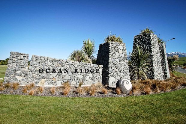 Exterior landscape entrance feature with a low, rugged grey stone wall and two taller stone pillars bearing the words “OCEAN RIDGE.” Sculptural stonework, yucca-like plants, ornamental grasses, a manicured lawn, clear blue sky, and distant mountainous terrain create a resort or residential gateway setting.