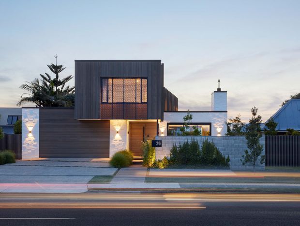 Street-facing exterior view of a contemporary multi-level residence at dusk. The home features a dark vertically ribbed upper facade, a large illuminated patterned window, a recessed timber entry, a broad integrated garage, pale masonry walls, low landscaped planting, and a flat-roof composition with a prominent chimney. Long-exposure vehicle light trails run across the foreground street.