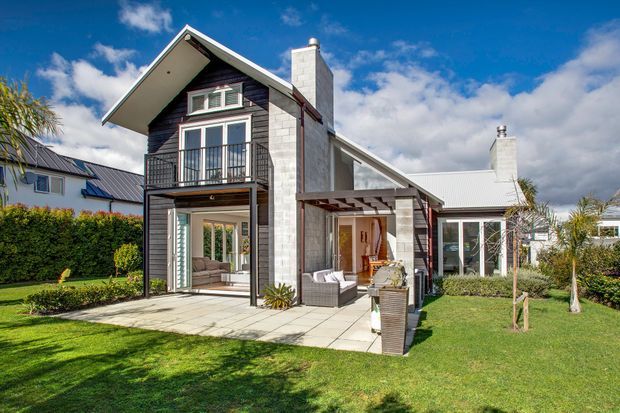 Exterior view of a contemporary multi-level home with a steep gabled roof, dark vertical cladding, light masonry chimney walls, large glazed openings, upper balcony, and covered outdoor patio. The landscaped garden and bright blue sky surround the house.