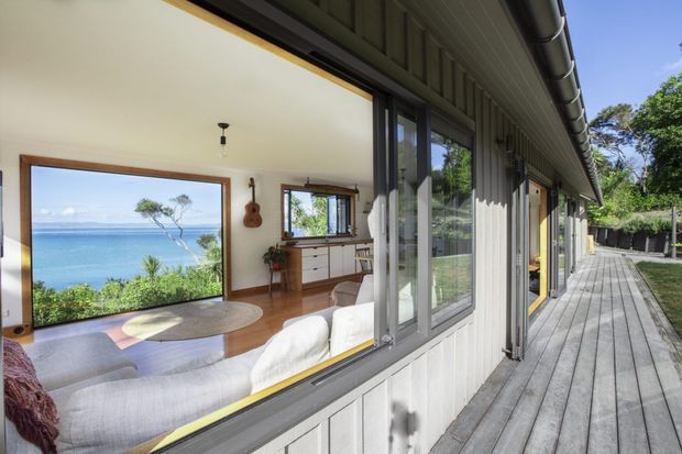 Exterior view along a timber-clad residential wing with extensive glazing opening onto a wide timber deck. Through the windows, a bright living room with pale seating, timber flooring, a console, and warm wood-framed openings is visible, with a blue coastal outlook beyond.