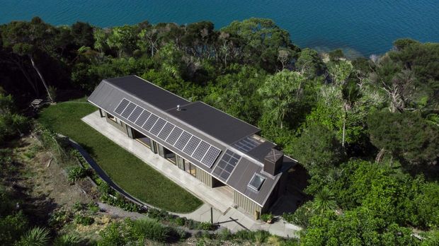 Aerial view of a long, low-profile contemporary house nestled among dense coastal vegetation beside blue water. The dark pitched roof has extensive integrated solar panels and skylights, while the building is accessed by a curved path and surrounded by landscaped grounds.