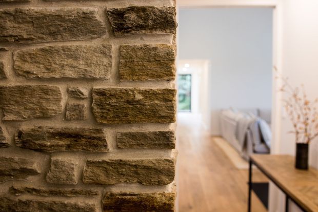 Interior view featuring a prominent rough-textured exposed stone or masonry wall in the foreground, opening toward a bright, narrow living or circulation space with warm timber flooring, a simple timber console table, black metal framing, neutral furnishings, and a small indoor tree. The image has shallow depth of field and a warm, contemporary rustic atmosphere.