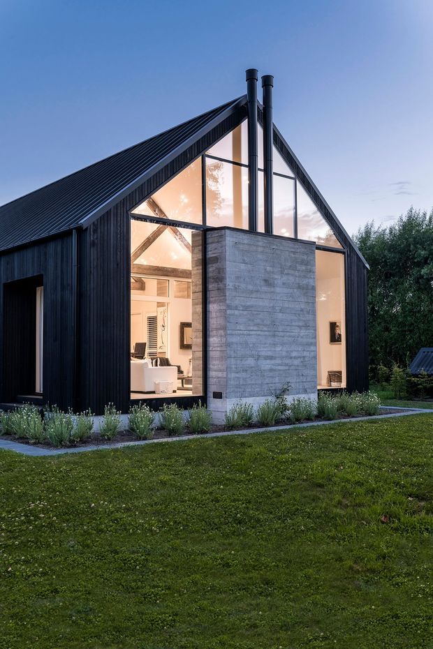 Exterior view of a contemporary gable-roof house at dusk, with dramatic floor-to-ceiling glazed end walls, black vertical cladding and roofing, a projecting grey concrete chimney volume with twin metal flues, and warm-lit living spaces visible inside. The house is set within a landscaped grassy garden with planted borders.