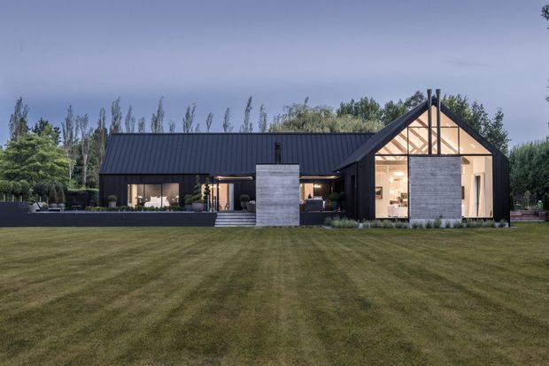 A contemporary single-storey house viewed from across a broad manicured lawn. The dark metal-clad building has a long pitched roof, a prominent glazed gable end with exposed timber-like structural framing, and contrasting light grey masonry feature walls. The surrounding landscape includes mature trees, clipped planting, and a low outdoor terrace.
