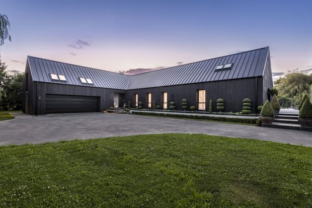 A substantial single-storey modern residence with a dramatic gabled metal roof and dark vertical cladding. The L-shaped facade includes a wide integrated garage, narrow vertical windows and doors, manicured topiary landscaping, a broad paved forecourt, and a large lawn. The image is an exterior architectural photograph taken at dusk.