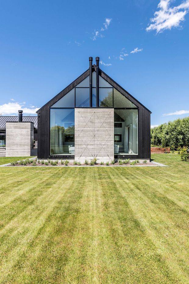 Front exterior view of a contemporary gable-roofed house set behind a broad manicured lawn. The facade combines dark vertical cladding, extensive floor-to-ceiling glazing, a central pale concrete or rendered feature wall, and twin black flues rising above the roof. A low adjoining wing, planted border, timber fencing, and clear blue sky complete the scene.